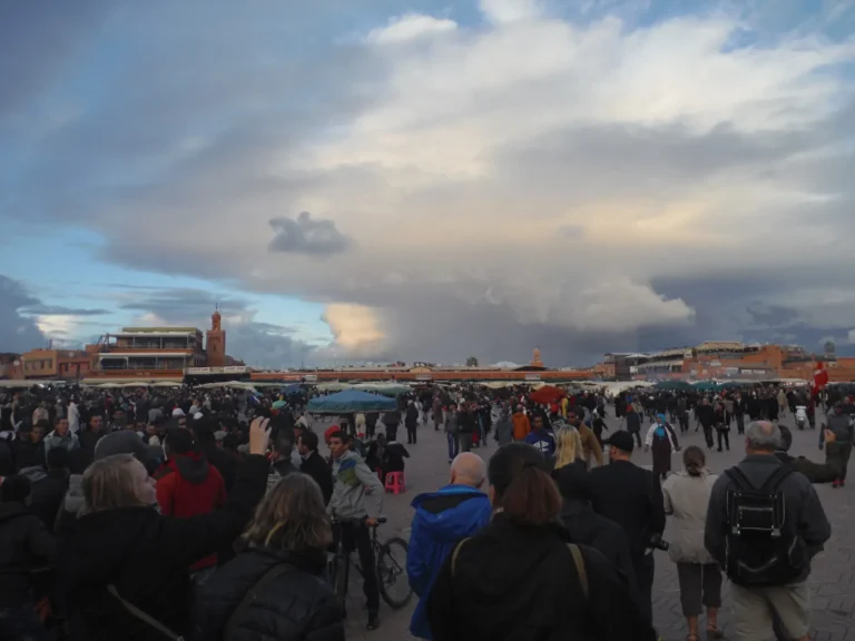 Marrakech in May: Is It Worth Visiting? 1 Djemaa el-Fna square in Marrakech at dusk, crowds filling the wide square with food stall tents and a minaret visible against a dramatic cloudy sky