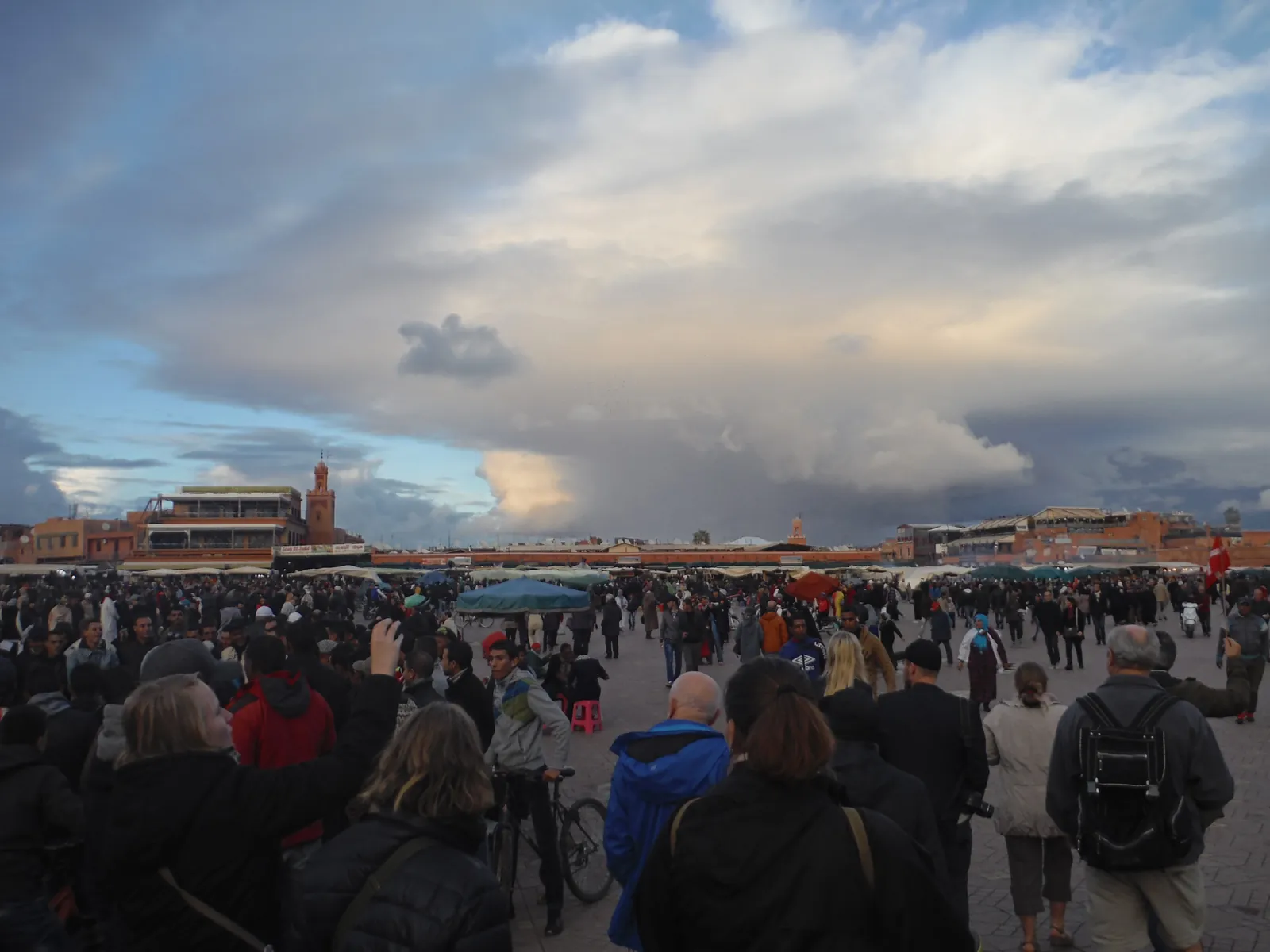 Djemaa el-Fna square in Marrakech at dusk, crowds filling the wide square with food stall tents and a minaret visible against a dramatic cloudy sky