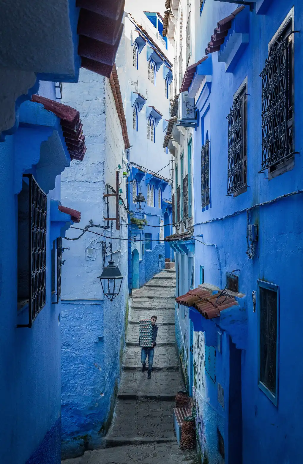 Man carrying a tray up a narrow stepped lane between blue houses in Chefchaouen